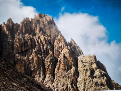 Die zerklüfteten Felsgipfel der Dolomitenklassiker ragen steil in den strahlend blauen Himmel, und weiße Wolkenfetzen verdecken teilweise die Langkofelscharte. Die schroffe Struktur der Felsen wird durch das Sonnenlicht wunderschön hervorgehoben.