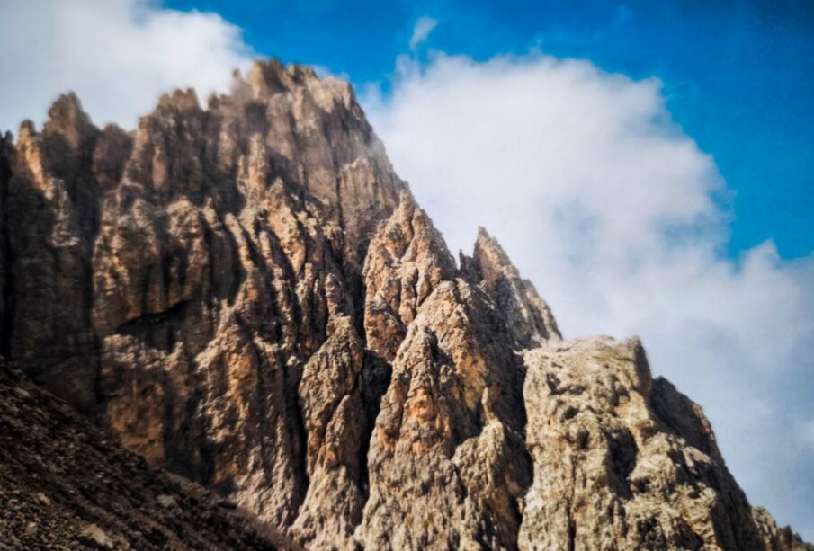 Die zerklüfteten Felsgipfel der Dolomitenklassiker ragen steil in den strahlend blauen Himmel, und weiße Wolkenfetzen verdecken teilweise die Langkofelscharte. Die schroffe Struktur der Felsen wird durch das Sonnenlicht wunderschön hervorgehoben.