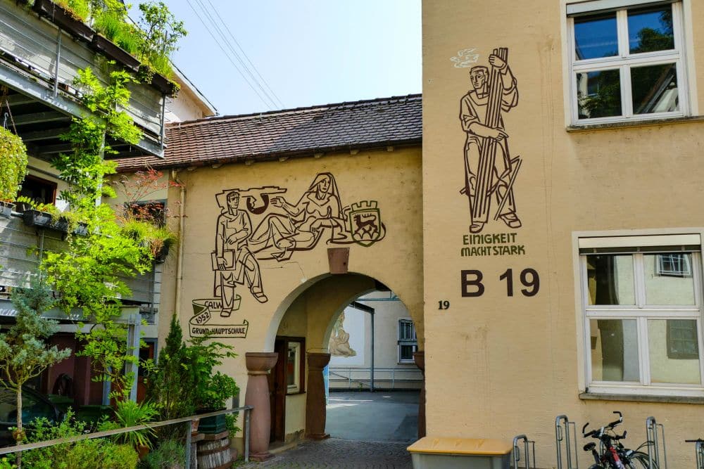 Ein beigefarbenes Gebäude in Calw mit Line-Art-Wandmalereien: eine zeigt drei Figuren und ein Wappen über einem Torbogen, die andere eine stehende Figur, die einen Stab hält, daneben steht "EINIGKEIT MACHT STARK". Der Innenhof ist mit Pflanzen und Fahrrädern bepflanzt.