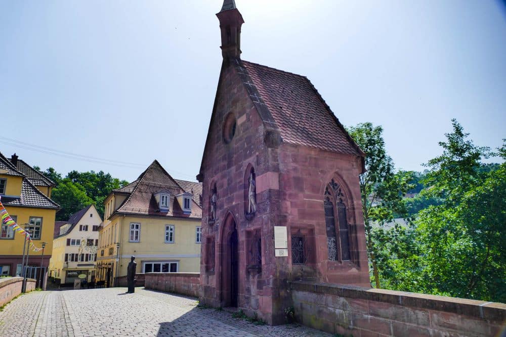 Eine kleine Steinkapelle mit Fenstern im gotischen Stil steht an einer Kopfsteinpflasterstraße in Calw, umgeben von gelben Gebäuden und Grünpflanzen unter einem klaren blauen Himmel.