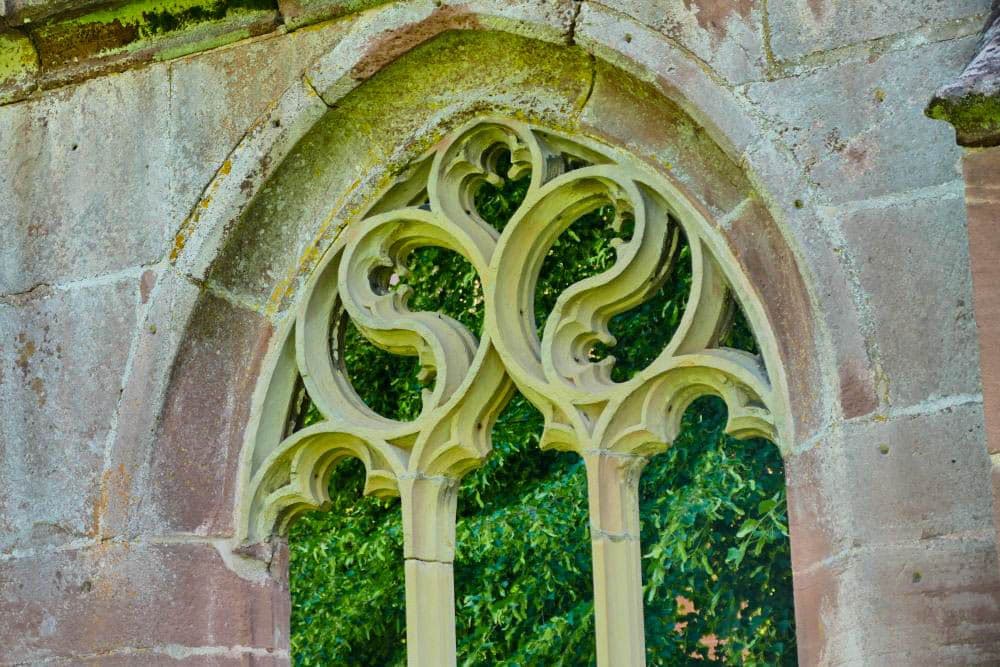 Nahaufnahme eines alten steinernen gotischen Fensters mit kunstvollem Maßwerk in Hirsau, das in eine verwitterte Mauer eingelassen ist, wobei durch die Fensteröffnungen grünes Laub zu sehen ist.