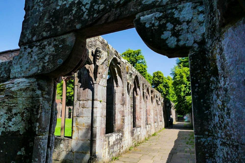 Alte Steinruinen von Hirsau mit gewölbten Fenstern und verwitterten Mauern, teilweise mit Moos bedeckt, säumen einen sonnenbeschienenen, von grünen Bäumen gesäumten Weg unter einem klaren blauen Himmel.