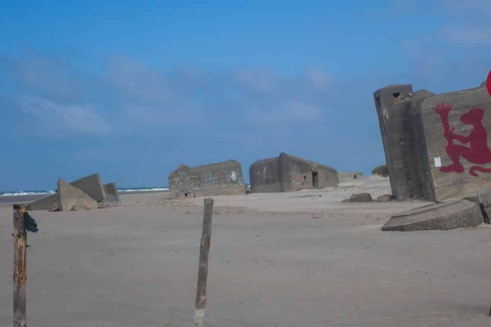 Betonbunker, verstreut über einen Sandstrand in Blavand, Dänemarks Westen, unter blauem Himmel. Auf einen Bunker ist ein roter Löwe gemalt. Im Hintergrund ist das Meer zu sehen, und im Vordergrund stehen Holzpfähle.