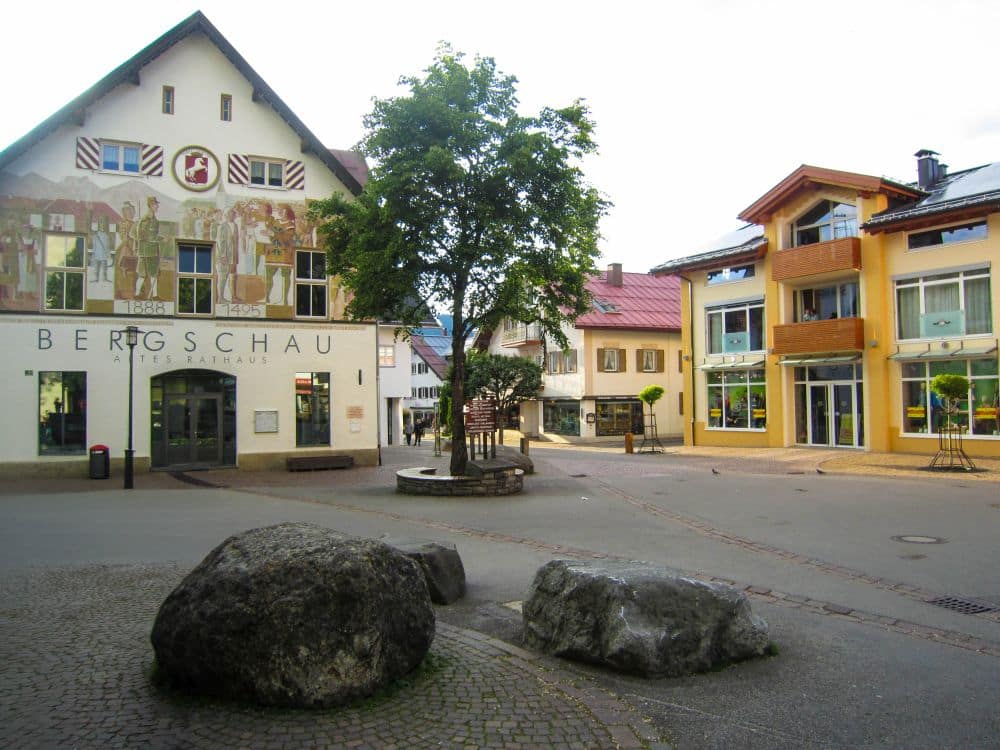 Ein kleiner europäischer Stadtplatz in Oberstdorf mit einem Baum in der Mitte, großen Steinen entlang des Kopfsteinpflasters und bunten Gebäuden mit Wandmalereien und Balkonen. Die friedliche Alpenlandschaft wirkt ruhig und beschaulich.