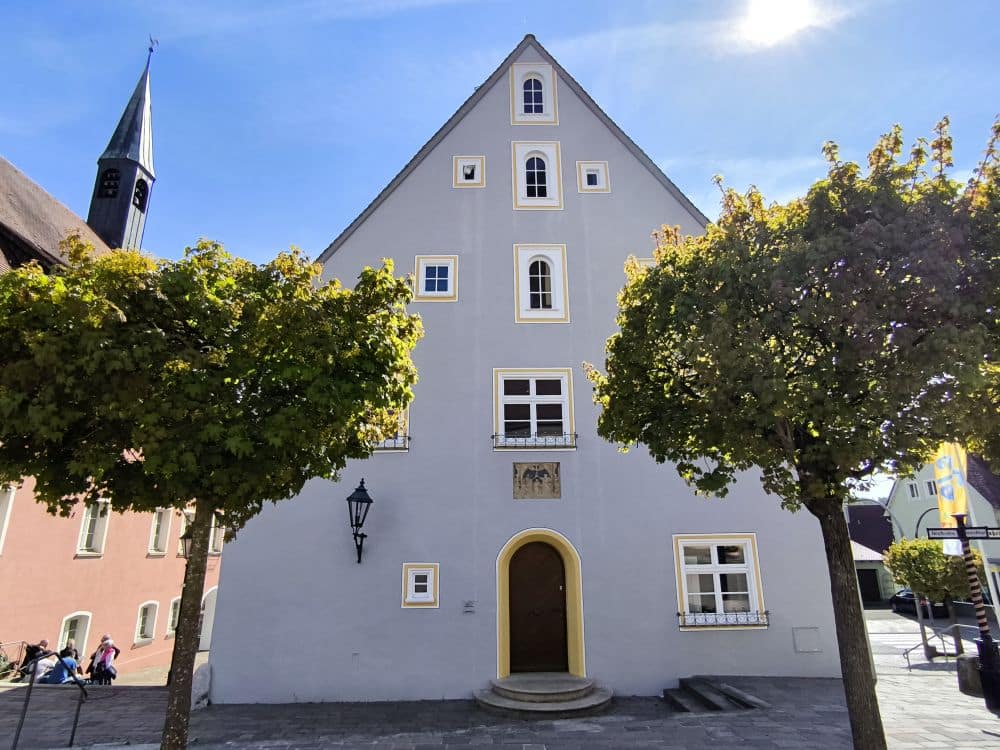 Ein hohes, hellgraues, dreieckiges Gebäude mit weißen und gelben Verzierungen, einer gewölbten Holztür und mehreren Fenstern steht zwischen zwei belaubten Bäumen unter einem klaren blauen Himmel in der Klosterstadt Neresheim. Im Hintergrund links erhebt sich ein Kirchturm.