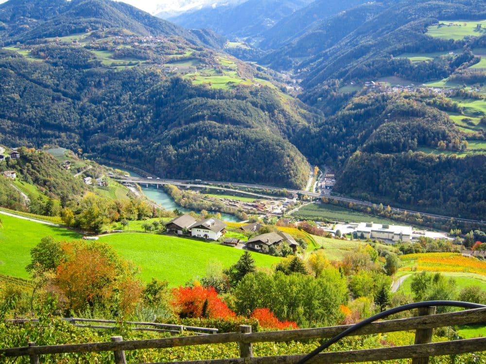 Ein malerisches Tal in Feldthurns, Südtirol, mit grünen Hügeln, bunten Herbstbäumen, verstreuten Häusern, einem gewundenen Fluss und fernen Bergen unter einem strahlenden Himmel. Ein Holzzaun säumt den Vordergrund entlang des berühmten Keschtnwegs.