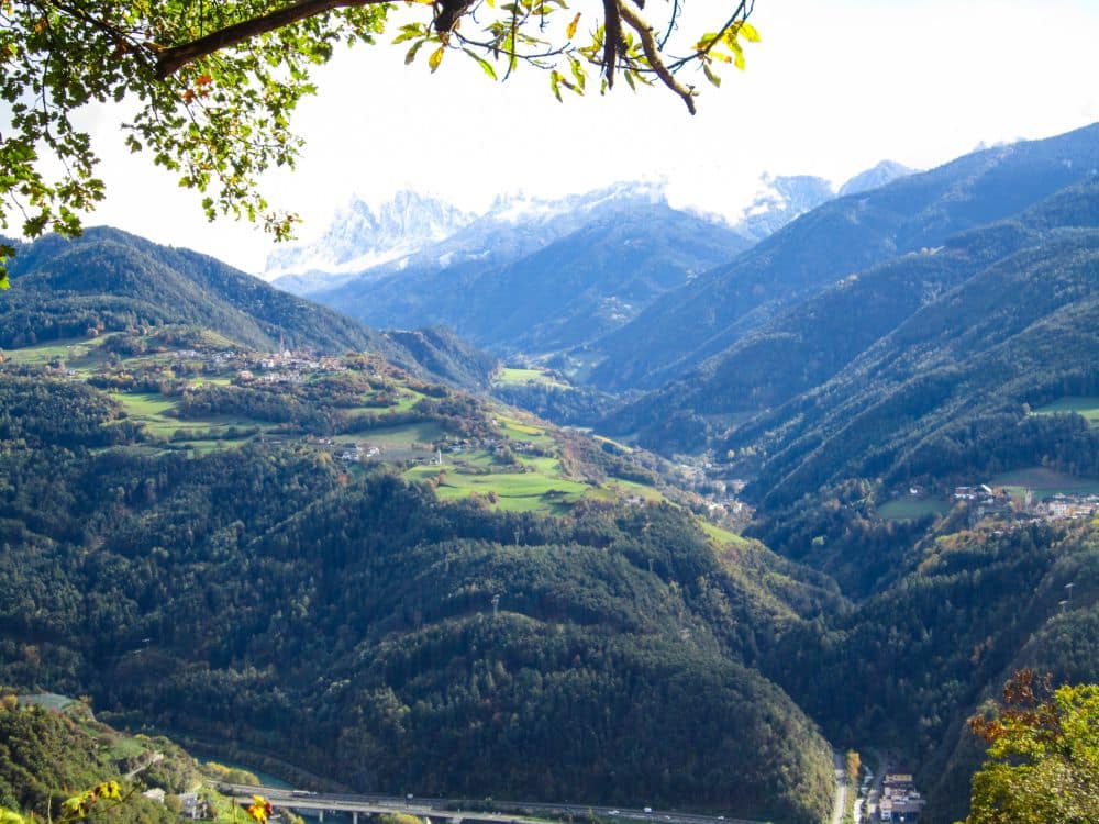 Blick auf ein üppig grünes Tal bei Feldthurns in Südtirol, umgeben von bewaldeten Hügeln und fernen schneebedeckten Bergen unter einem klaren Himmel, mit verstreuten Gebäuden und Baumzweigen, die den oberen Teil des malerischen Keschtnwegs einrahmen.