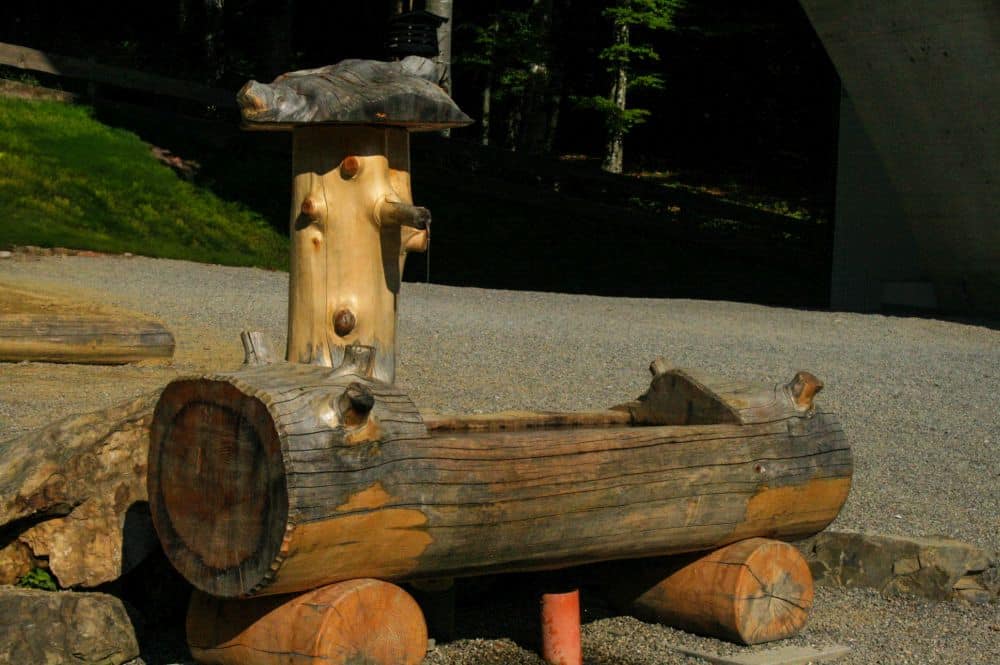Eine rustikale Holzbank aus großen Stämmen mit natürlichen Ästen und Zweigen, die im Freien auf Kies mit Grünpflanzen und Felsen im Hintergrund steht, erinnert an den Charme eines Alpenrefugiums bei Oberstdorf.
