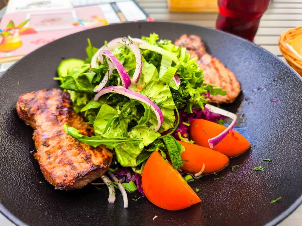 Auf einem Tisch in Calw steht ein schwarzer Teller mit gegrilltem Fleisch, frischem grünen Salat, geschnittenen roten Zwiebeln, Gurken, Tomatenscheiben und zerkleinertem violettem Kraut, im Hintergrund sind ein Getränk und die Speisekarte zu sehen.