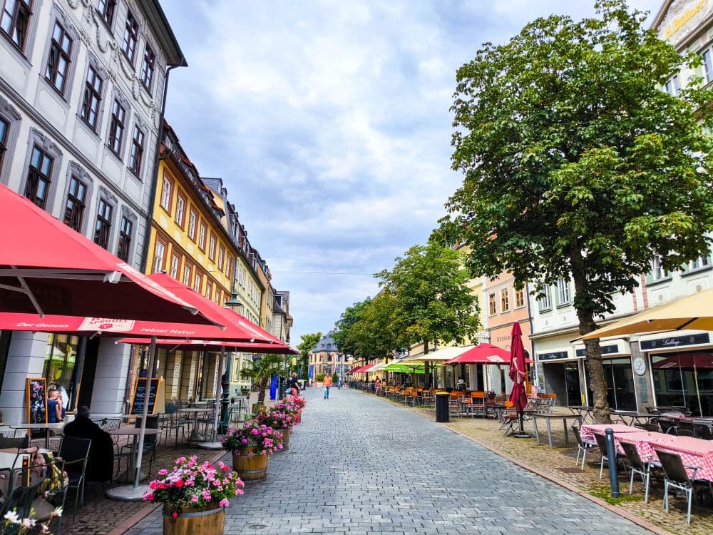 Eine charmante Kopfsteinpflasterstraße in Fulda, gesäumt von bunten Gebäuden, Straßencafés, roten Sonnenschirmen und Blumenkübeln, mit grünen Bäumen auf beiden Seiten unter einem teilweise bewölkten Himmel.