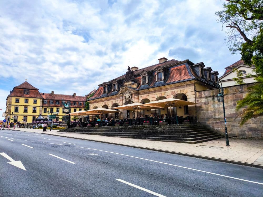 Ein historisches europäisches Gebäude in Fulda mit Sitzplätzen im Freien unter großen Sonnenschirmen säumt eine breite, leere Straße an einem bewölkten Tag. Die Menschen essen draußen und genießen die klassische Architektur im Hintergrund.