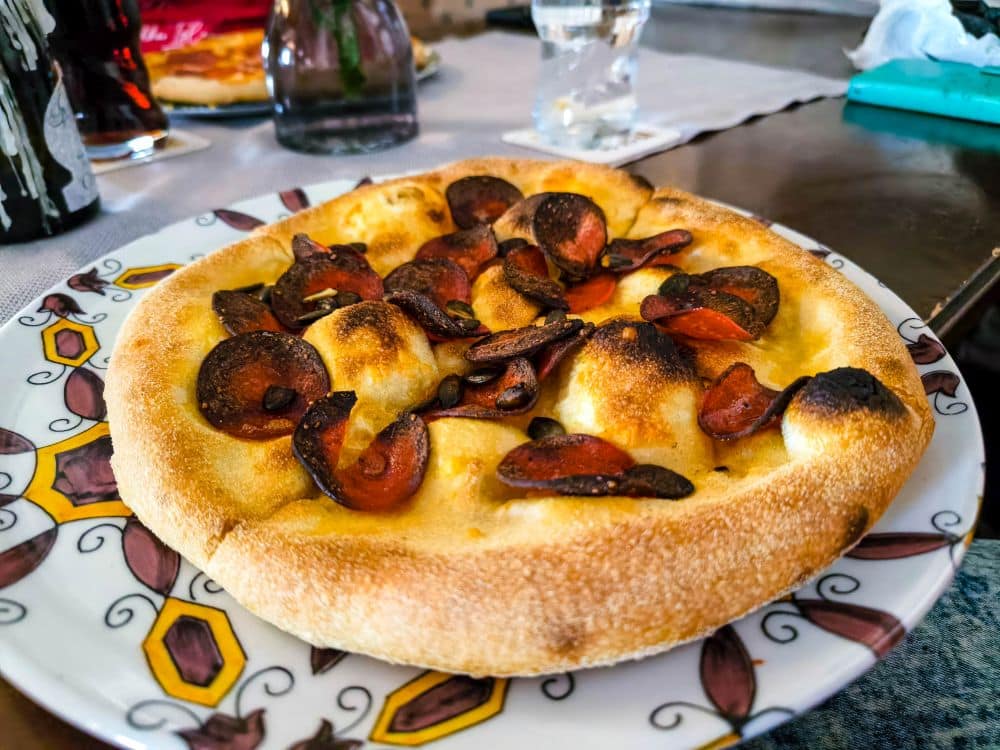 Eine kleine Peperoni-Pizza mit einer dicken, goldenen Kruste und knusprigen, leicht verkohlten Peperoni liegt auf einem dekorativen Teller in Fulda, mit Getränken und unscharfen Gegenständen im Hintergrund.