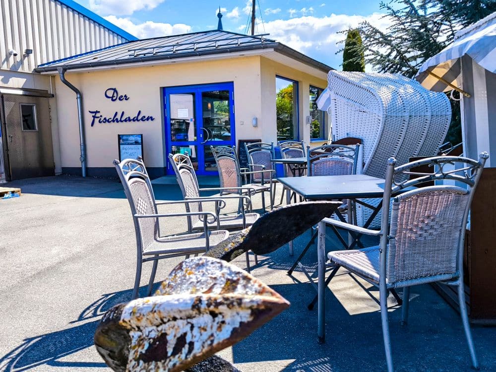 Freisitz mit Metallstühlen und -tischen vor dem Fischladen in Kühlungsborn, mit blau gerahmten Türen und Fenstern. Ein dekorativer Anker bildet den Rahmen für eine perfekte Ostsee-Auszeit unter freiem Himmel.