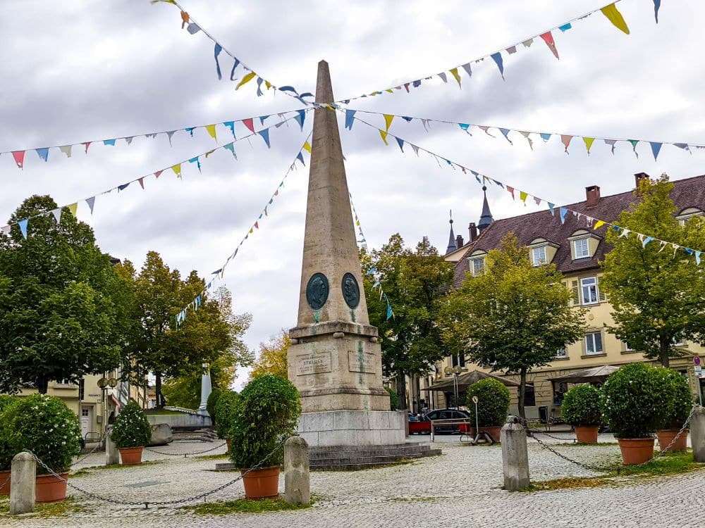 Auf einem kopfsteingepflasterten Platz in Ludwigsburg steht ein steinernes Obeliskdenkmal, das mit bunten dreieckigen Wimpeln geschmückt ist. Grüne Bäume, bunte Gärten und Topfpflanzen umgeben es, im Hintergrund stehen die Gebäude der Barocke Pracht unter einem wolkenverhangenen Himmel.