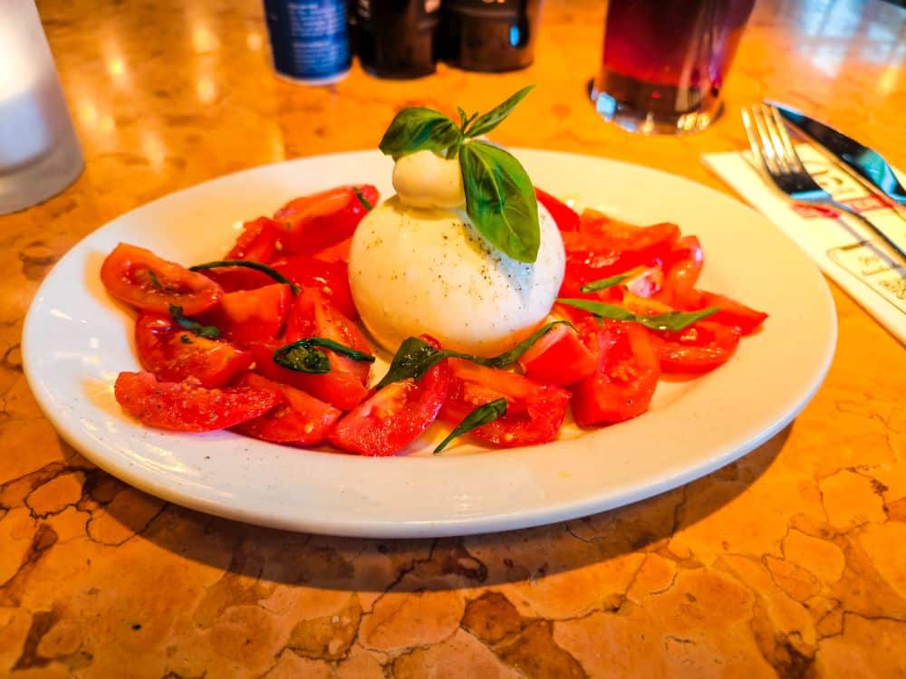 Ein Teller mit Caprese-Salat mit geschnittenen Tomaten, frischen Basilikumblättern und einer Kugel Burrata-Käse in der Mitte, garniert mit Pfeffer und Basilikum, serviert auf einem Marmortisch, der an die Eleganz der Ludwigsburger Barockgärten erinnert.