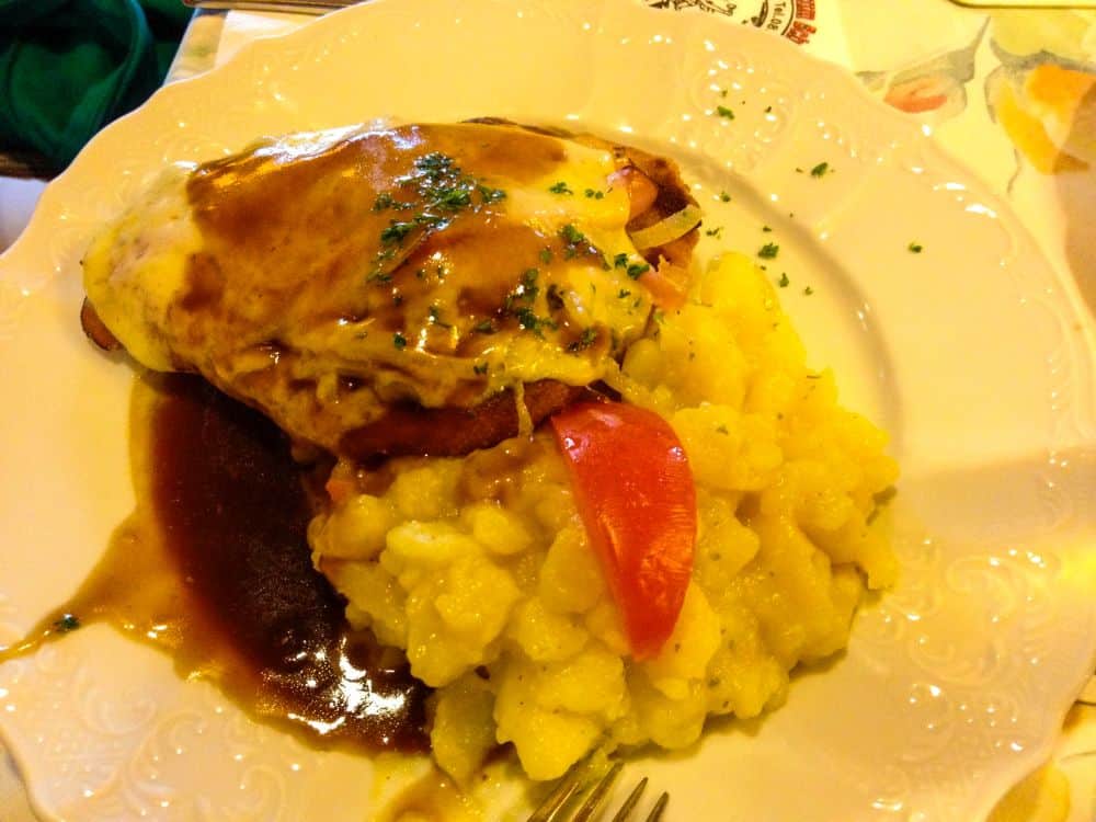 Ein Teller mit einer Portion Fleisch, überzogen mit geschmolzenem Käse und brauner Bratensoße, garniert mit Kräutern, erinnert an die deftigen Aromen der Alpen bei Oberstdorf, serviert mit gelbem Kartoffelsalat und einer frischen Tomatenscheibe.