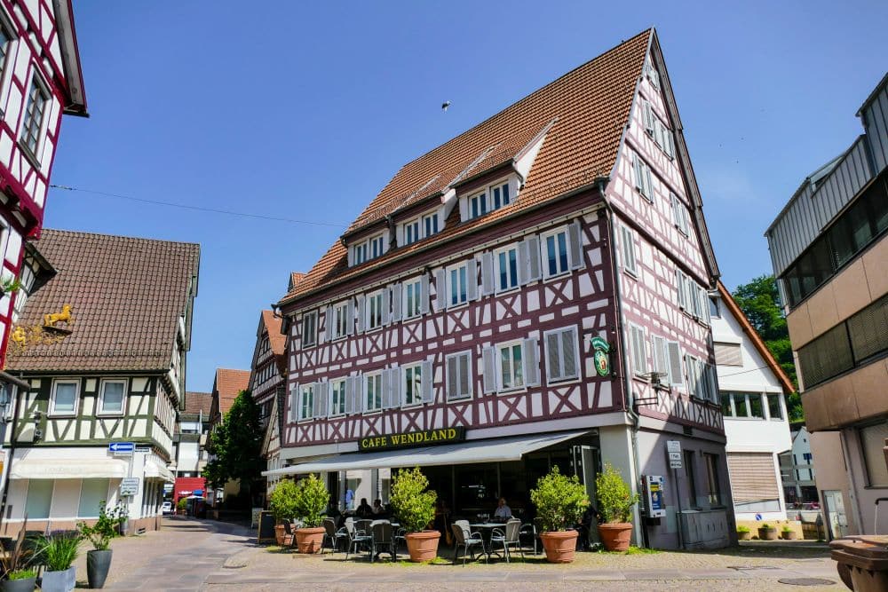 Ein traditionelles Fachwerkhaus mit steilem Dach beherbergt das Café Wendland in Calw, umgeben von Tischen im Freien, Topfpflanzen und benachbarten historischen Gebäuden unter einem strahlend blauen Himmel.