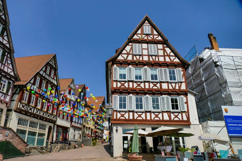 Eine Straßenszene in Calw zeigt bunte Luftballons, die zwischen traditionellen Fachwerkhäusern unter blauem Himmel aufgereiht sind; ein Gebäude ist wegen Renovierungsarbeiten teilweise abgedeckt. Im Vordergrund sind Sitzgelegenheiten im Freien mit Sonnenschirmen zu sehen.