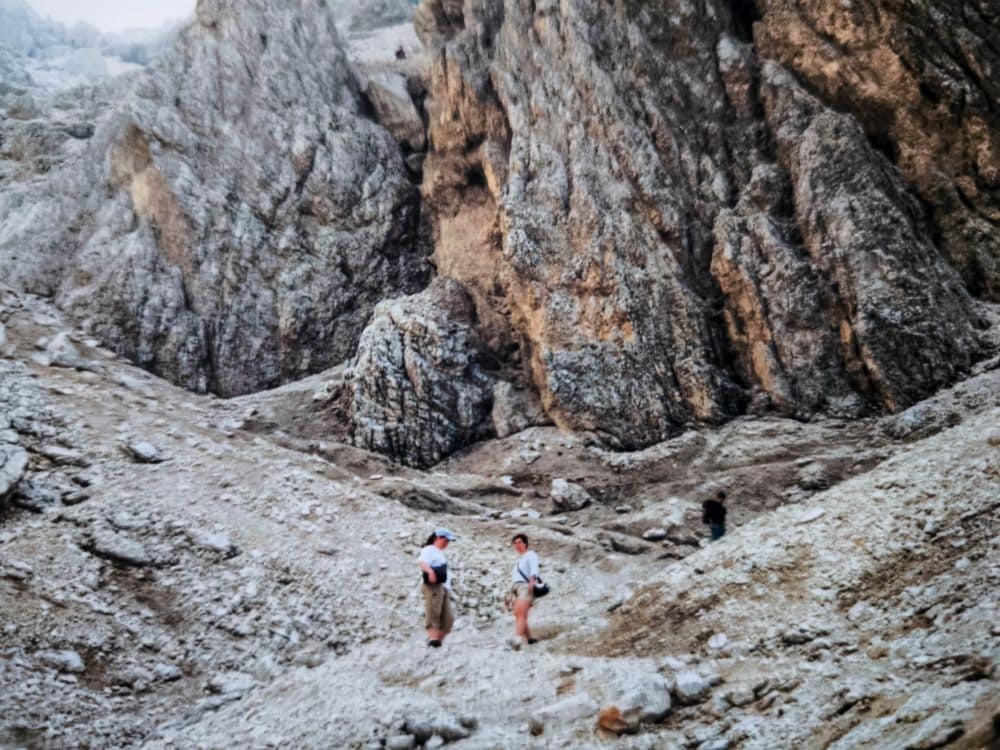 Zwei Wanderer mit kurzen Hosen und Rucksäcken stehen an einem felsigen Dolomitenklassiker-Hang, hinter dem sich bei bedecktem Himmel große zerklüftete Felsformationen erheben.