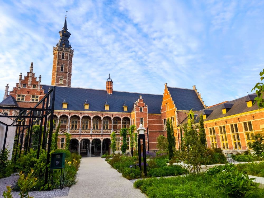 Ein historisches Backsteingebäude mit einem hohen Uhrenturm und Bogenfenstern, dieses Kleinod lässt Sie charmant übernachten im Herzen der flämischen Stadt, umgeben von einem gepflegten Garten und Wegen unter einem blauen Himmel.