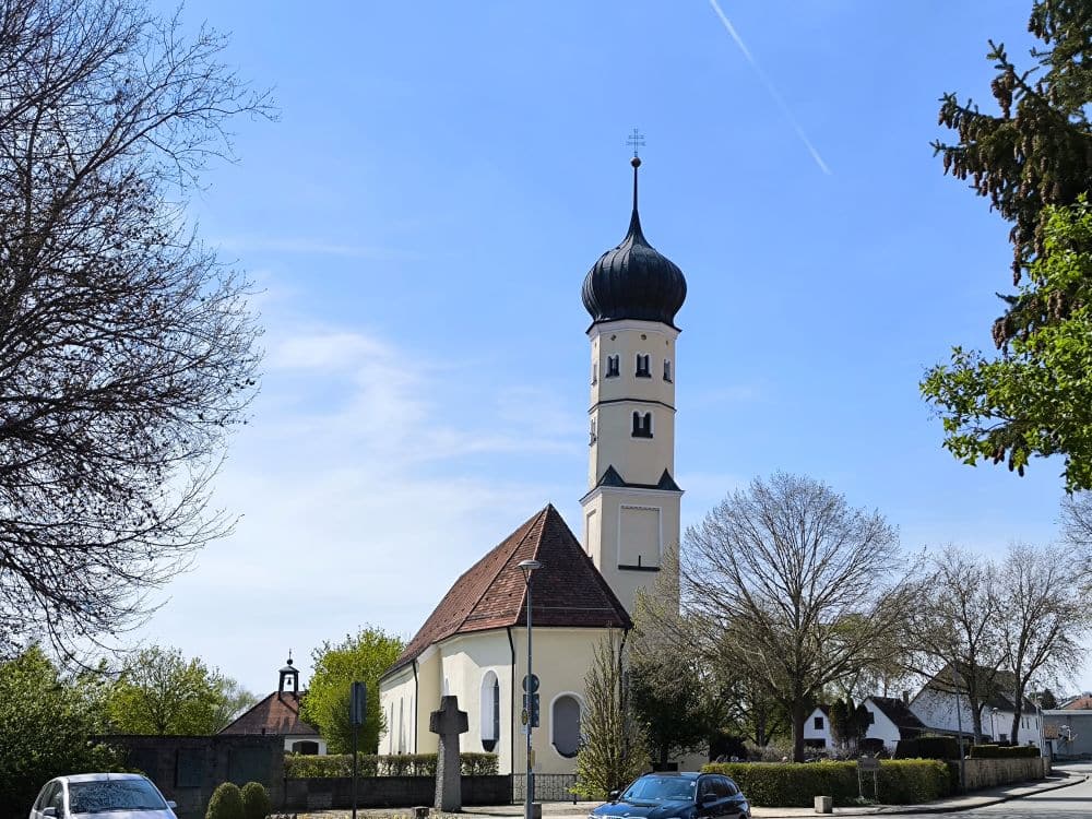 In Neresheim, der Klosterstadt, steht zwischen kahlen Bäumen unter strahlend blauem Himmel eine weiße Kirche mit einem hohen, runden, zwiebelförmigen Kuppelturm. In der Nähe sind Autos geparkt, und vor dem historischen Kloster steht ein Steinkreuz.