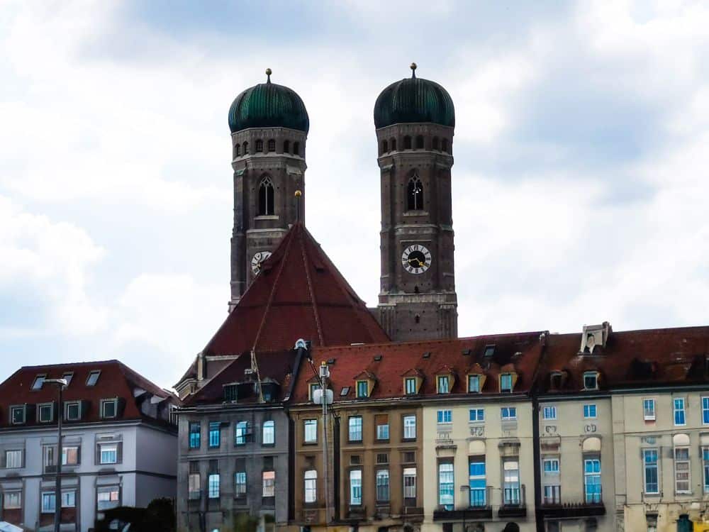 Das Bild zeigt die beiden grünen Kuppeltürme der Frauenkirche, die sich über bunten, historischen Gebäuden in München, Deutschland, unter einem teilweise bewölkten Himmel erheben.