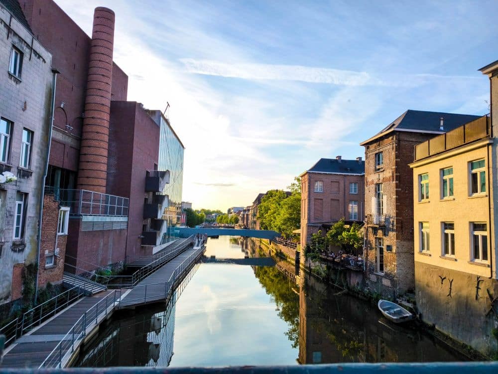 Ein ruhiger Kanal verläuft zwischen Backsteingebäuden mit einem Gehweg auf der linken Seite. Bäume säumen das rechte Ufer, an dem ein kleines Boot vertäut ist. Entdecken Sie Hotels in Mechelen in der Nähe für charmante Übernachtungen in dieser flämischen Stadt, die sich im stillen Wasser spiegelt.