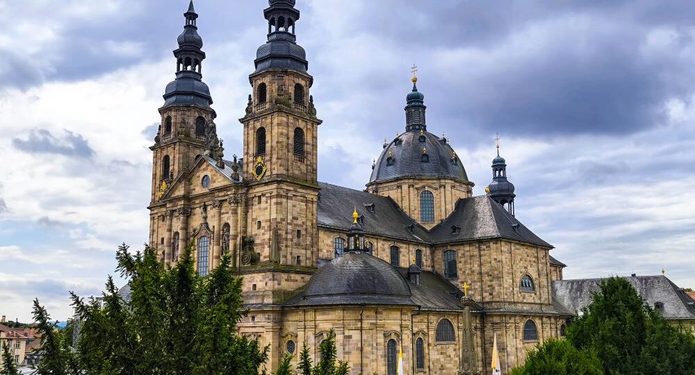 Der große barocke Fuldaer Dom mit zwei hohen Türmen und einer zentralen Kuppel aus hellem Stein hebt sich deutlich von einem wolkenverhangenen Himmel ab. Üppige grüne Bäume umgeben das Gebäude.