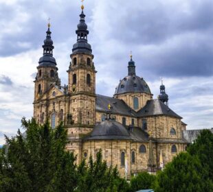 Der große barocke Fuldaer Dom mit zwei hohen Türmen und einer zentralen Kuppel aus hellem Stein hebt sich deutlich von einem wolkenverhangenen Himmel ab. Üppige grüne Bäume umgeben das Gebäude.