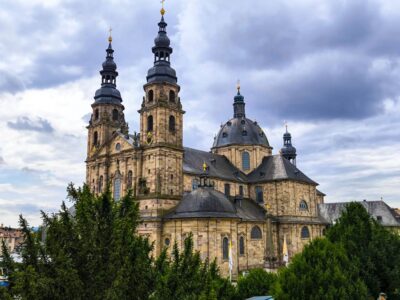 Der große barocke Fuldaer Dom mit zwei hohen Türmen und einer zentralen Kuppel aus hellem Stein hebt sich deutlich von einem wolkenverhangenen Himmel ab. Üppige grüne Bäume umgeben das Gebäude.