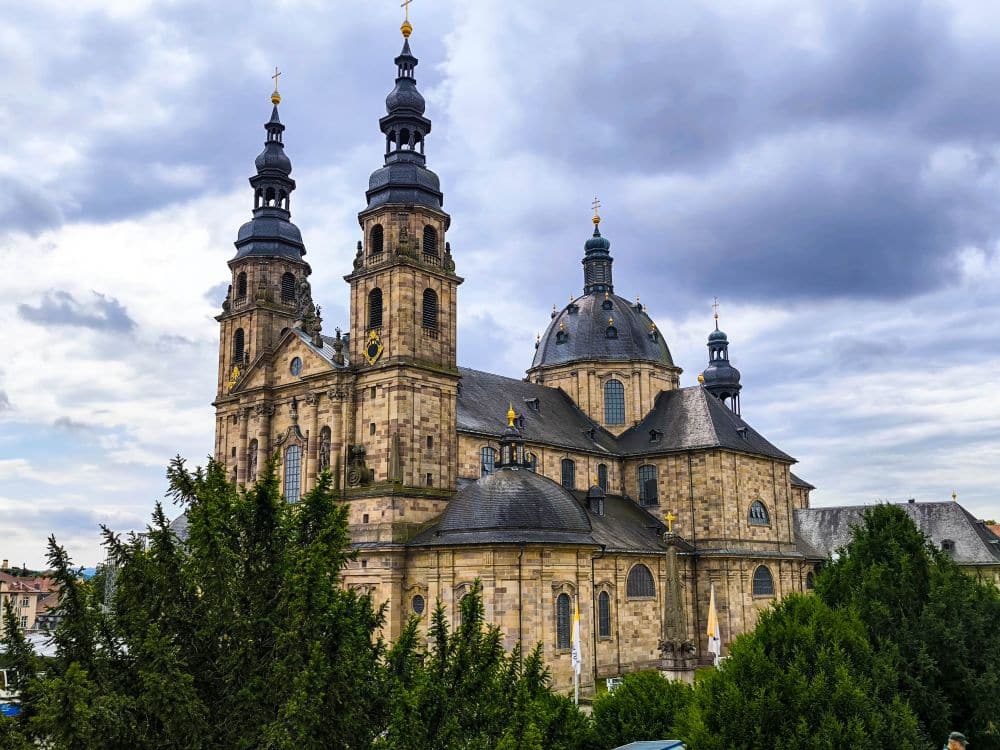 Der große barocke Fuldaer Dom mit zwei hohen Türmen und einer zentralen Kuppel aus hellem Stein hebt sich deutlich von einem wolkenverhangenen Himmel ab. Üppige grüne Bäume umgeben das Gebäude.