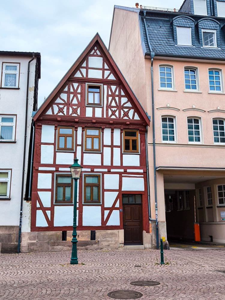 Ein traditionelles Fachwerkhaus mit weißen Wänden und roten Holzbalken steht zwischen zwei modernen Gebäuden an einer Kopfsteinpflasterstraße in Fulda, mit einer grünen Straßenlaterne davor.