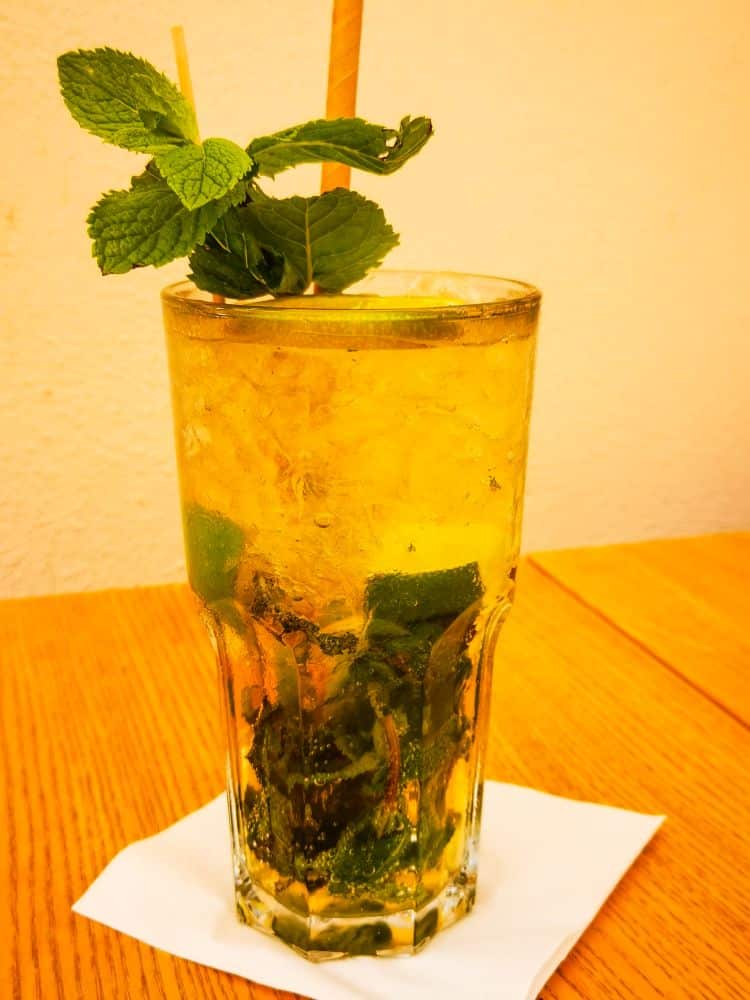 Ein hohes Glas eisgekühlter Mojito, garniert mit frischen Minzblättern und einem Strohhalm, steht auf einer weißen Serviette auf einem Holztisch und erinnert vor einem hellen Hintergrund an die entspannte Erfurter Café-Kultur.