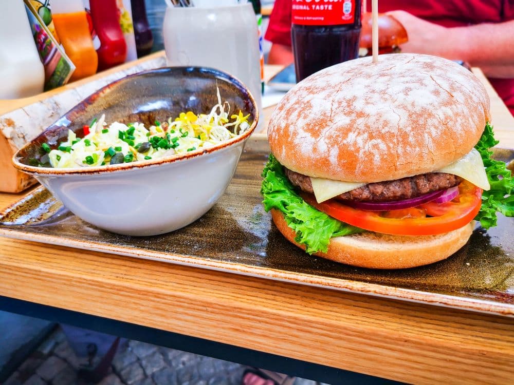 Ein Cheeseburger mit Salat, Tomate, Zwiebel und Käse im Brötchen liegt auf einem Teller neben einer Schüssel Salat mit Mais, Erbsen und Sprossen auf einem Holztisch in Erfurt.