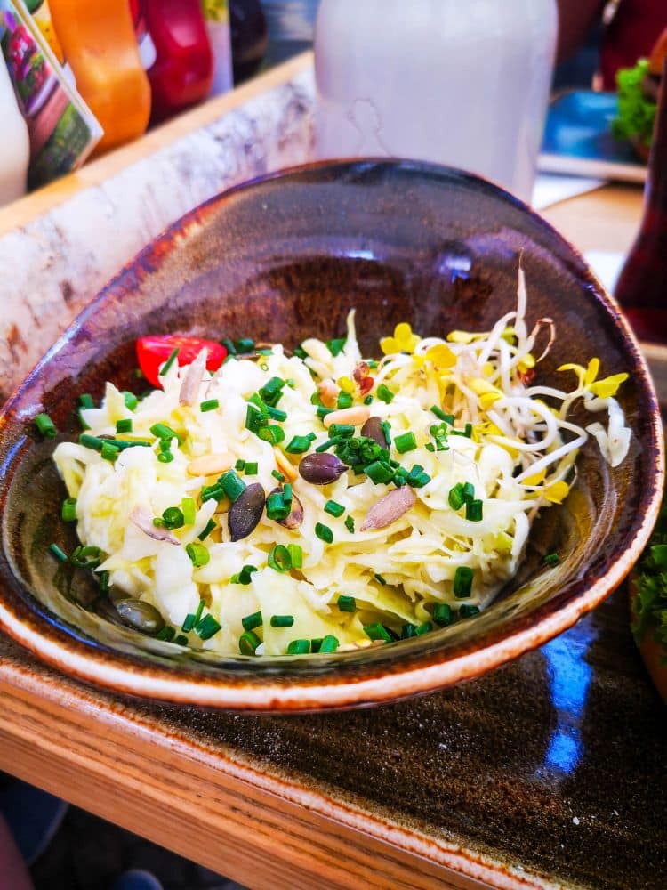 Eine rustikale braune Schüssel mit einem frischen Krautsalat, der mit Schnittlauch, Sprossen, Samen und ein paar Tomatenstücken gefüllt ist, steht auf einem Holztisch in Erfurt, mit Gewürzen und Flaschen im verschwommenen Hintergrund.