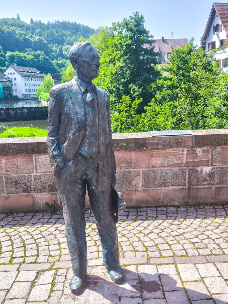 Die Bronzestatue eines Mannes im Anzug und mit Hut steht auf einem Kopfsteinpflasterweg in der Nähe einer Steinmauer in Calw, mit grünen Bäumen, Gebäuden und bewaldeten Hügeln im Hintergrund an einem sonnigen Tag.