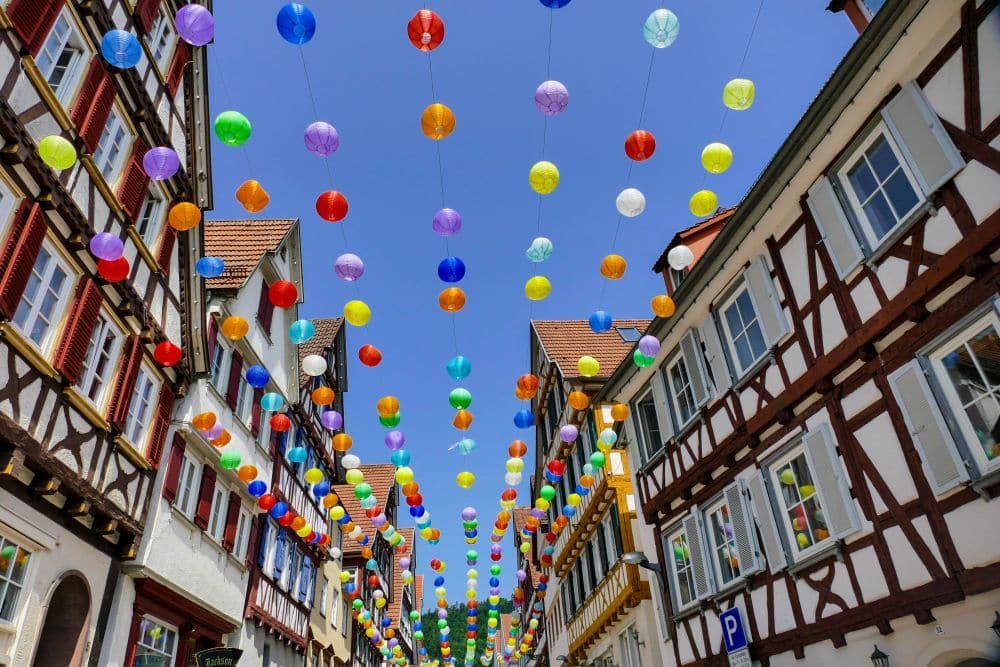 Bunte Papierlaternen hängen über einer schmalen Straße in Calw, die von traditionellen Fachwerkhäusern gesäumt ist, unter einem klaren blauen Himmel. Die festliche Dekoration schafft eine lebendige und fröhliche Atmosphäre.