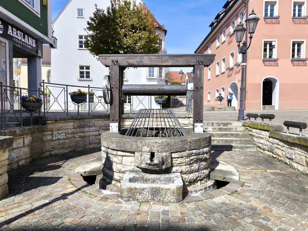 Ein steinerner Brunnen mit einer Holzkonstruktion steht auf einem gepflasterten Platz in der Klosterstadt Neresheim, umgeben von Bänken, Blumenkästen und farbenfrohen Gebäuden unter einem klaren blauen Himmel.