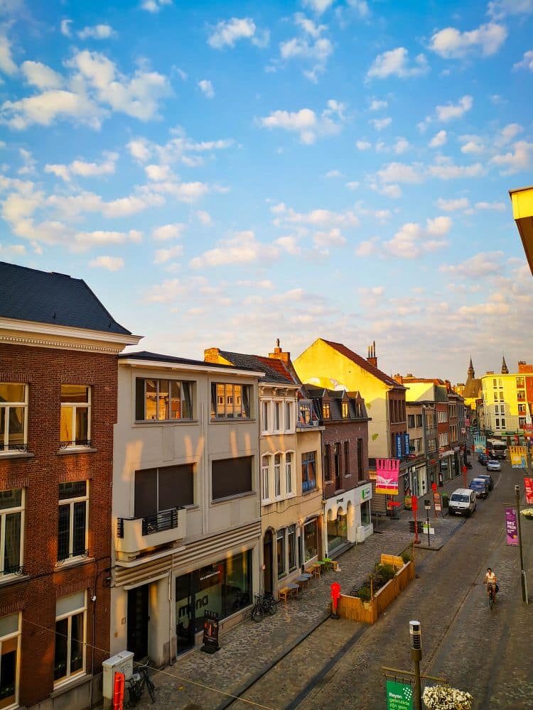 Eine lebendige Straßenszene in einer flämischen Stadt mit farbenfrohen Gebäuden, Fahrrädern auf dem Bürgersteig und ein paar Autos auf der Kopfsteinpflasterstraße unter blauem Himmel - perfekt für charmante Übernachtungen in lokalen Hotels.