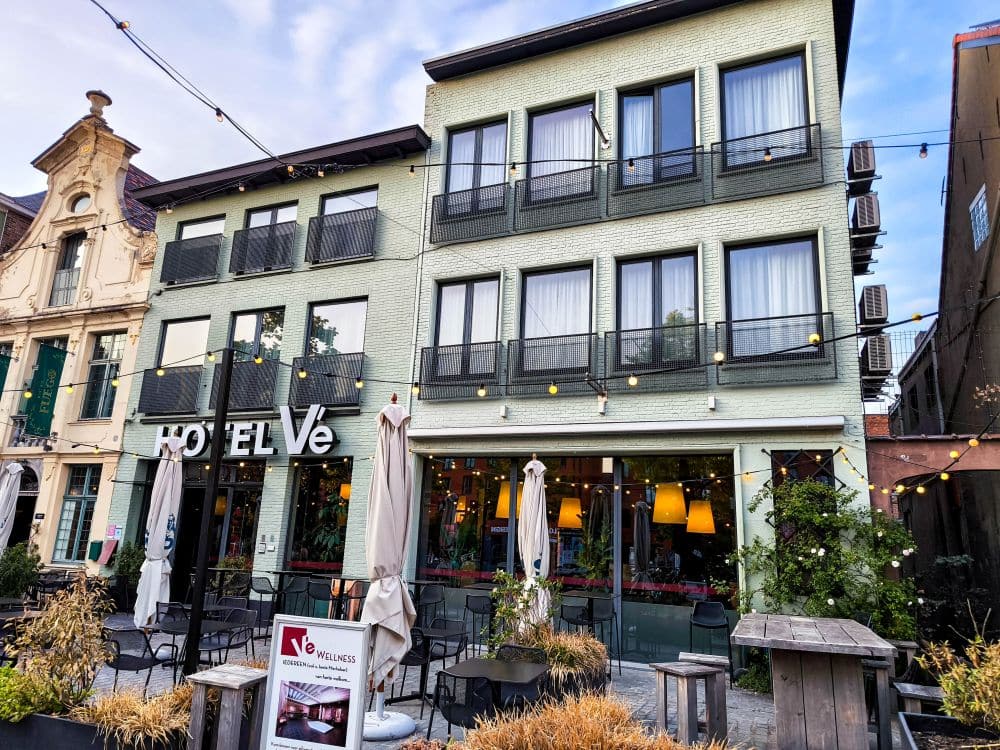 Ein modernes dreistöckiges Hotel mit großen Fenstern und Balkonen, Sitzgelegenheiten im Freien mit Tischen und Sonnenschirmen, Lichterketten und einem Schild mit der Aufschrift Hotel Vé - ein einladender Ort, um in der charmanten flämischen Stadt Mechelen zu übernachten.