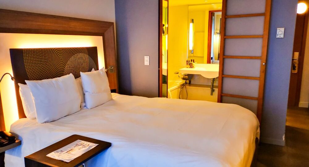 Ein ordentlich gemachtes Hotelbett mit weißer Bettwäsche und Kissen steht neben einem kleinen Tisch. Im Hintergrund ist durch eine hölzerne Schiebetür ein modernes Badezimmer mit heller Beleuchtung zu sehen - perfekt für entspannende Aufenthalte in der flämischen Stadt Mechelen.