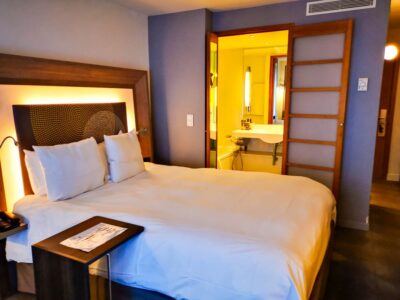 Ein ordentlich gemachtes Hotelbett mit weißer Bettwäsche und Kissen steht neben einem kleinen Tisch. Im Hintergrund ist durch eine hölzerne Schiebetür ein modernes Badezimmer mit heller Beleuchtung zu sehen - perfekt für entspannende Aufenthalte in der flämischen Stadt Mechelen.