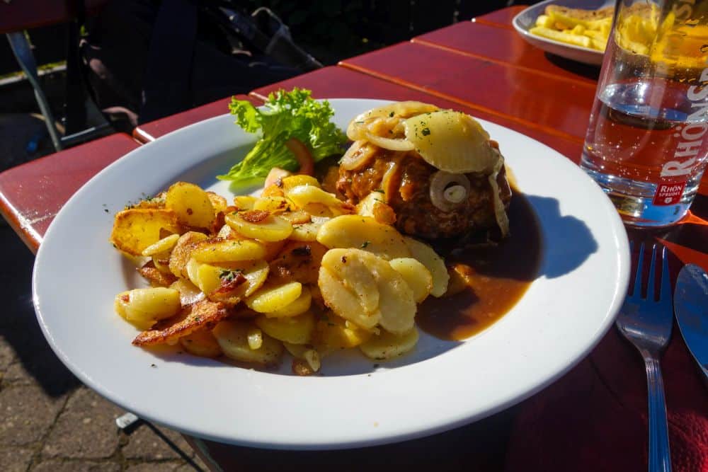 Ein weißer Teller mit gebratenen Kartoffelscheiben, einer Fleischpastete mit gekochten Zwiebeln und brauner Bratensoße, dazu eine kleine Salatbeilage - perfekt nach einem Rhöner Wanderabenteuer. Ein Glas Wasser und eine Gabel stehen neben dem Teller auf einem roten Tisch.