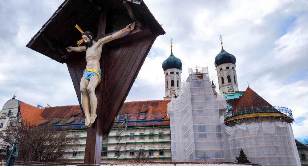 Ein großes Kruzifix steht auf einem Friedhof mit Grabsteinen, vor der Barockkirche Kloster Benediktbeuern mit zwei Zwiebeltürmen. Ein Teil des Gebäudes ist wegen Renovierungsarbeiten mit Gerüsten und Netzen verhüllt, vor einem wolkenverhangenen Himmel und fernen Bergen.