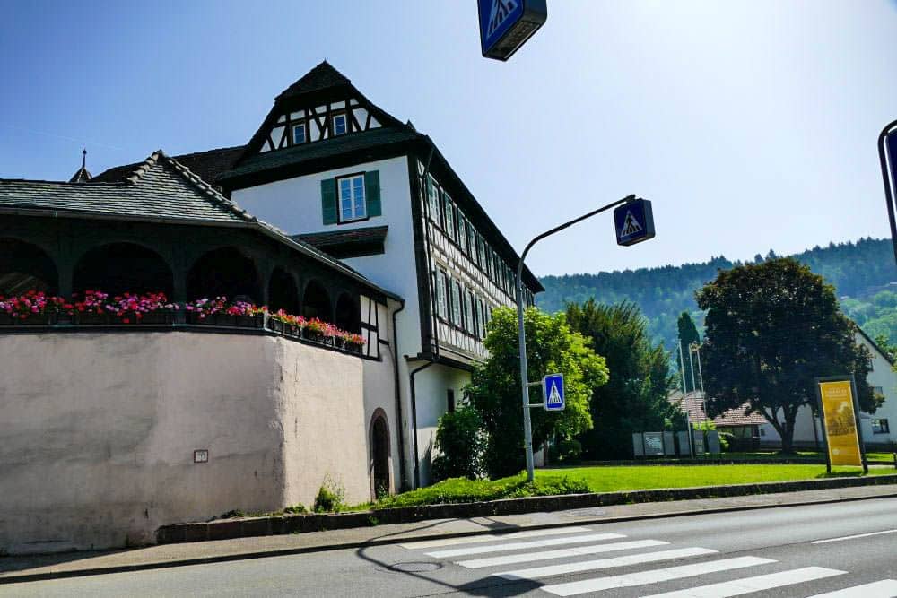 Ein Fachwerkhaus mit grünen Fensterläden steht an einer Straße in Hirsau, in der Nähe eines Fußgängerüberwegs und von Straßenschildern. Blumenkästen säumen den Balkon, und im Hintergrund sind bei klarem Himmel Bäume und Hügel zu sehen.