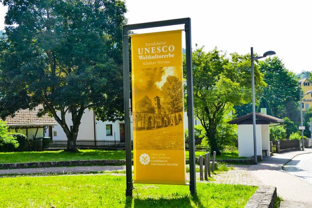 Ein gelbes Schild mit Informationen über Hirsau, das zum UNESCO-Welterbe gehört, steht an einem sonnigen Tag auf dem Bürgersteig, mit Bäumen und Wohnhäusern im Hintergrund.