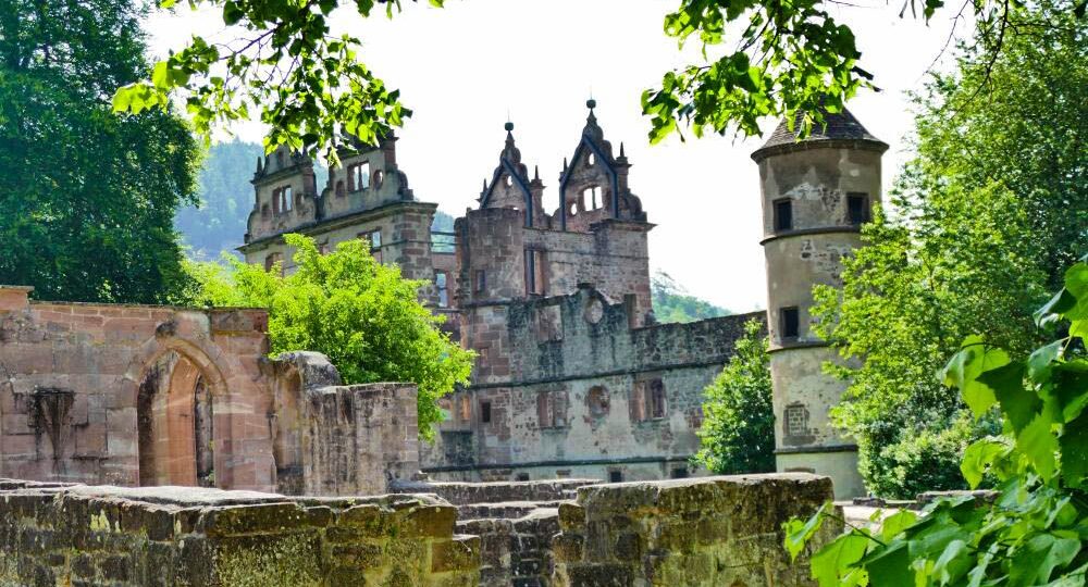 Ein historisches Steinschloss in Hirsau mit verschnörkelten Türmen und Bogenfenstern wird an einem sonnigen Tag teilweise von grünen Bäumen verdeckt und von alten Steinmauern umgeben.
