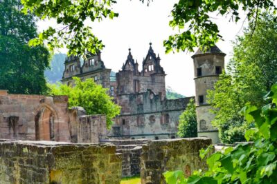 Ein historisches Steinschloss in Hirsau mit verschnörkelten Türmen und Bogenfenstern wird an einem sonnigen Tag teilweise von grünen Bäumen verdeckt und von alten Steinmauern umgeben.