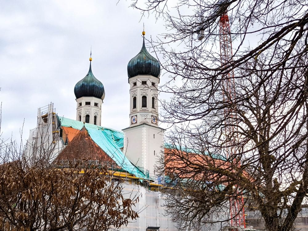 Eine Kirche mit zwei zwiebelförmigen Kuppeln, typisch für die Barockarchitektur, ist teilweise von Gerüsten und Planen verdeckt. Laublose Bäume rahmen die Szene ein, und im Hintergrund steht ein roter Kran, der auf die Restaurierungsarbeiten im Kloster Benediktbeuern hinweist.