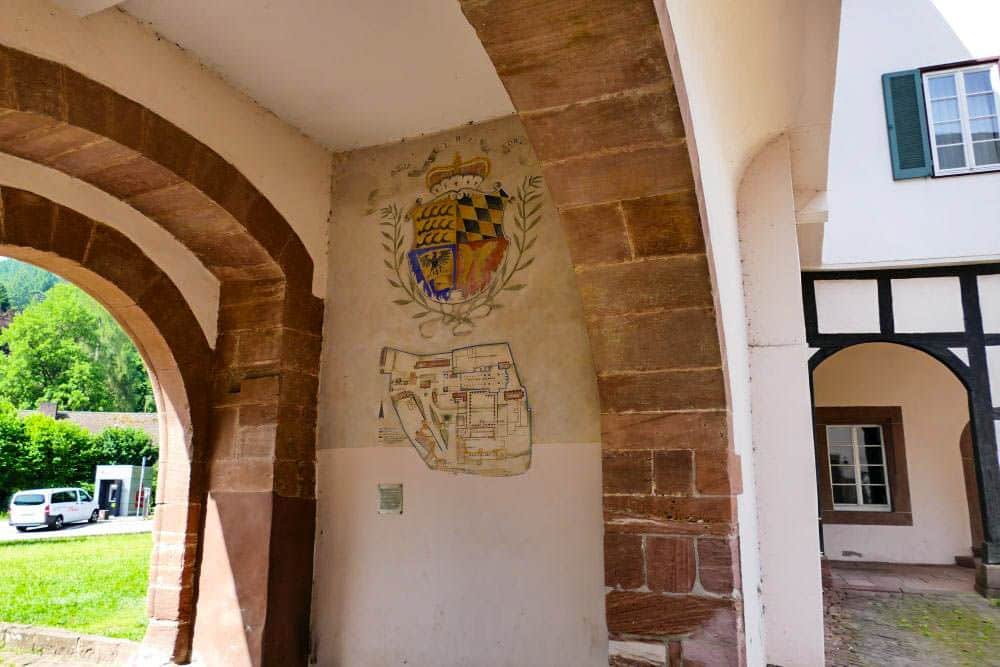 Ein gewölbter Eingang aus Stein in Hirsau zeigt ein gemaltes Wappen und eine alte Landkarte an der Wand. Das Sonnenlicht erhellt den Raum und gibt den Blick frei auf einen grasbewachsenen Hof, geparkte Autos und ein Gebäude, das direkt vor der Tür steht.